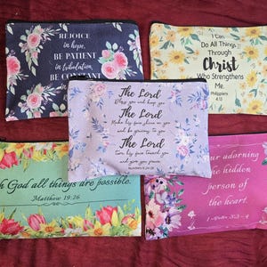 May include: Five rectangular zippered pouches featuring floral patterns and religious text. The pouches are navy blue, yellow, light purple, mint green, and pink. Each displays a unique scripture verse printed in black, decorative script and serif fonts.