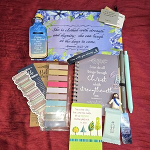 May include: A collection of religious stationery items including a floral zippered pouch with Proverbs 31:25-26, a spiral notebook with Philippians 4:13, a chapstick holder, colorful sticky tabs, pens, a small Jesus figurine, and various scripture cards.