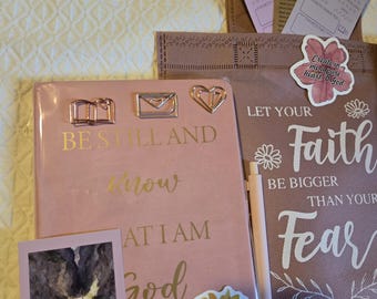 May include: A pink journal with gold text reading BE STILL AND KNOW THAT I AM GOD, featuring three metal paperclips, a sunflower sticker, and a small card. Beside it, a brown bag displays the text LET YOUR FAITH BE BIGGER THAN YOUR FEAR.