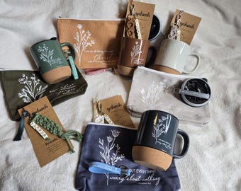 May include: A collection of ceramic mugs with cork bases, fabric zipper pouches, and macrame keychains. Each item features white floral line art and printed religious text. The mugs are in green, brown, gray, and black, while the pouches are tan, olive, gray, and navy blue.