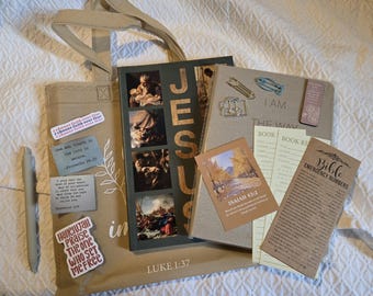 May include: A beige canvas tote bag featuring the text Luke 1:37 and various stickers, a book titled Jesus with religious imagery, a grey journal with the text I Am The Way, a pen, book review cards, a bookmark, and a Bible emergency numbers card.
