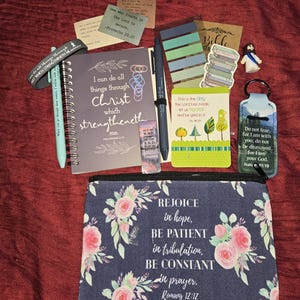 May include: A collection of religious stationery items including a spiral-bound notebook with the text I can do all things through Christ which strengtheneth me, a floral zippered pouch with Romans 12:12, pens, bookmarks, stickers, and a keychain.