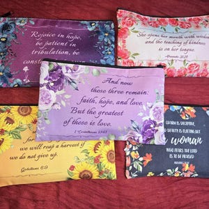 May include: Five rectangular zippered pouches featuring floral patterns and printed scripture verses. The designs include purple, pink, yellow, and dark backgrounds with various flower arrangements and text such as Rejoice in hope, She opens her mouth with wisdom, And now these three remain, we will reap a harvest, and Charm is deceptive.