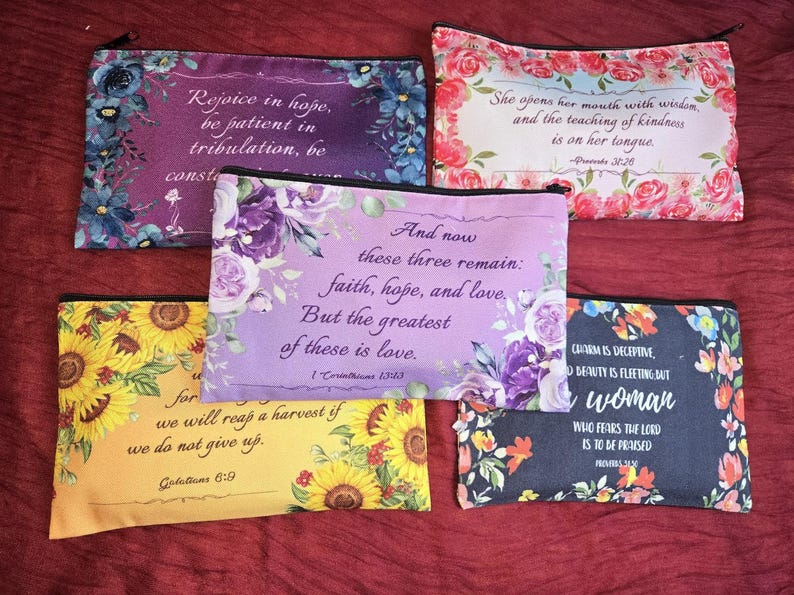May include: Five rectangular zippered pouches featuring floral patterns and printed scripture verses. The designs include purple, pink, yellow, and dark backgrounds with various flower arrangements and text such as Rejoice in hope, She opens her mouth with wisdom, And now these three remain, we will reap a harvest, and Charm is deceptive.