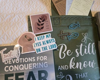 May include: A green fabric bag featuring the text Be still and know that, accompanied by a dragonfly graphic, two blue fish-shaped paper clips, a cross-shaped paper clip, and several religious stickers with text including Trust and I keep my eyes always on the Lord.