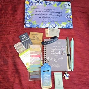 May include: A zippered pouch with blue floral patterns and the text She is clothed with strength and dignity, a spiral notebook with the text I can do all things through Christ, two pens, sticky note tabs, a keychain, and small religious items.