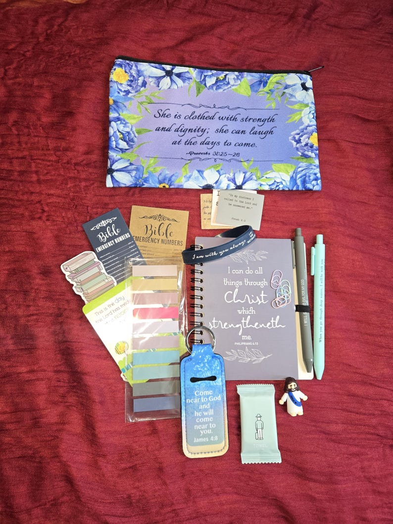 May include: A zippered pouch with blue floral patterns and the text She is clothed with strength and dignity, a spiral notebook with the text I can do all things through Christ, two pens, sticky note tabs, a keychain, and small religious items.