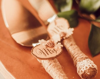 May include: A cake serving set featuring a metallic knife and server with handles wrapped in tan twine. Each handle is decorated with white lace trim, small fabric flowers, and a small wooden slice engraved with the words Mrs. and Mr.