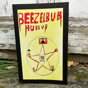 May include: A framed artwork with the words "BEEZELBUB HUBBUB" in red, stylized lettering. The artwork features a yellow background with a pentagram design and various illustrations. The artwork is displayed outdoors.