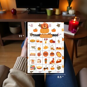 May include: Thanksgiving Bingo game, 8.5 x 11 inches, featuring a turkey and pumpkin design. The bingo card includes fall-themed illustrations such as pumpkins, pie, and leaves. The card is held by a person wearing a beige sweater and blue jeans.