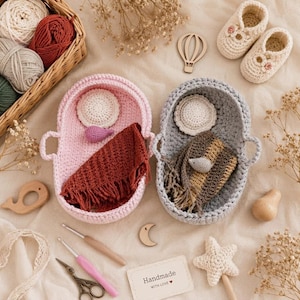 May include: Two oval crochet baskets in light pink and light gray, each containing a small round crocheted mat and a fringed blanket. The set includes cream-colored baby booties, wooden shapes, crochet hooks, scissors, and a card labeled Handmade.