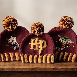 May include: Three maroon and gold knit beanies with pom-poms. The center beanie features the letters HP in gold. The left beanie displays an embroidered character with glasses and a scarf, while the right beanie shows a green creature with glasses.