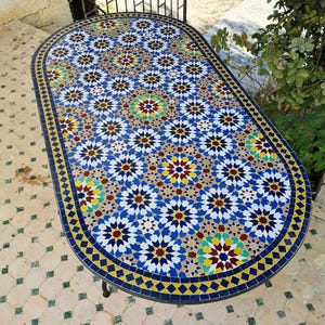 May include: An oval mosaic tile table with a colourful geometric pattern. The tabletop features a repeating design of blue, yellow, red, and white star-like shapes. The table has a dark metal frame and is set on a tiled surface.