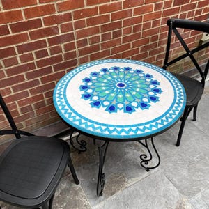 May include: A round bistro table with a mosaic tile top. The table features a geometric pattern in blue, turquoise, and white. It has a black metal frame and is paired with two black chairs. The table's diameter is approximately 75 cm.