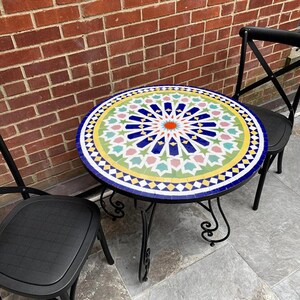 May include: A round mosaic tile table with a black metal frame. The tabletop features a colorful geometric pattern with blue, green, yellow, pink, and orange tiles. Two black chairs are positioned on either side of the table. The table is approximately 24 inches in diameter.