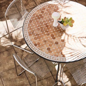 May include: A circular outdoor table with a mosaic tile surface, accompanied by three wire chairs. The table features a black border and a decorative design. A small white lamp and a potted succulent are on the table. The chairs are made of silver metal.