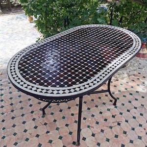 May include: Oval mosaic tile table with a black wrought iron base. The tabletop features a geometric pattern of dark brown and white tiles, framed by a white and black border. The table is outdoors on a tiled patio.