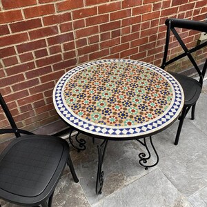 May include: A round mosaic tile table with a black metal frame, accompanied by two black chairs. The table's surface displays a pattern of blue, green, red, and brown stars. The table's diameter is approximately 76 cm.