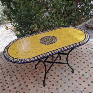May include: Oval mosaic tile table with a yellow and blue design. The tabletop features a yellow mosaic tile pattern with a blue and yellow border. The table has a black metal base.