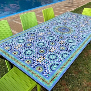 May include: A rectangular outdoor table with a mosaic tile top featuring a blue and white floral pattern. The table is surrounded by four lime green plastic chairs. The table's legs are black, and the setting is on a grassy area near a pool.