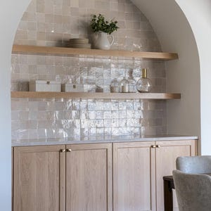 Puede incluir: Mueble de cocina empotrado con puertas y estantes de madera clara. El salpicadero presenta azulejos cuadrados y texturizados. Los elementos decorativos incluyen una jarra blanca con vegetación, platos y tarros de cristal.