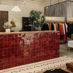 Puede incluir: Interior de una tienda con un mostrador con azulejos rojos y una encimera de madera. La ropa se exhibe en percheros. Una pequeña planta en maceta está sobre el mostrador. Una alfombra estampada está en primer plano.