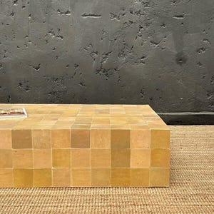 May include: A square coffee table made from small, square tiles in shades of beige and tan. The table is placed on a woven jute rug, with a dark grey textured wall behind it. The table's dimensions are approximately 100 cm x 100 cm.