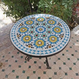 May include: Round mosaic tile table with a black metal frame. The tabletop features a geometric pattern in blue, yellow, white, and red tiles. The table has three curved legs and is suitable for outdoor use.