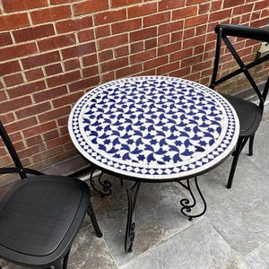 May include: A round bistro table with a mosaic tile top displaying a blue and white star design. The table has a black metal frame and is accompanied by two black chairs. The table's diameter is approximately 76 cm.