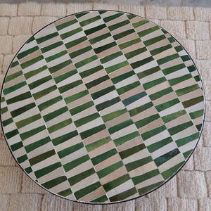 Op de afbeelding: Ronde tafel met een mozaïektegelblad. Het tegelpatroon heeft afwisselende rijen groene, beige en witte rechthoekige tegels. De tafel heeft een zwarte rand en staat op een neutraal gekleurd tapijt.