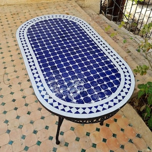 May include: Oval mosaic tile table with a blue and white geometric pattern. The tabletop features a grid of blue squares and a white border with a triangle design. The table has a black metal frame and legs.