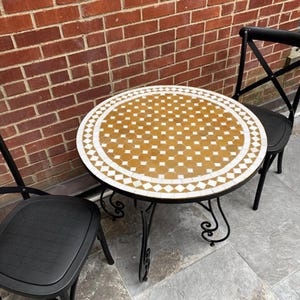 May include: A small outdoor bistro set featuring a round mosaic tile table and two black metal chairs. The table has a decorative pattern of yellow and white tiles. The chairs have a crisscross back design.