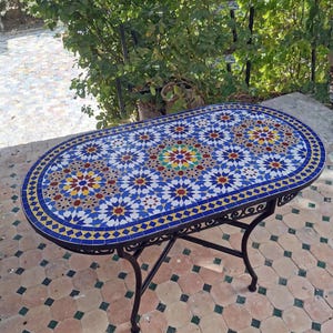 Op de afbeelding: Een ovale mozaïektafel met een zwart smeedijzeren onderstel. Het tafelblad heeft een kleurrijk geometrisch patroon in blauw, geel, rood en wit. De tafel is ongeveer 152 cm lang en 76 cm breed.