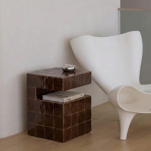 May include: A brown tiled side table with a silver bowl on top. The table has a unique design with an open shelf holding a book. A white chair is in the background, and the table is on a wooden floor.