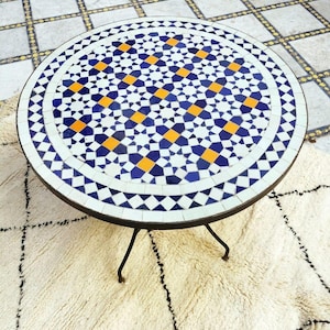 May include: Round mosaic tile table with a black metal frame and legs. The tabletop features a geometric pattern of blue, white, and orange tiles. The table is set on a cream-colored rug with a black geometric design.