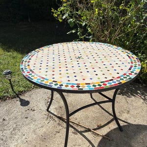 May include: Round outdoor mosaic table with a cream-colored surface and colorful square tiles. The table has a black metal frame and is approximately 36 inches in diameter.