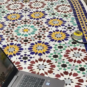 May include: A round mosaic tile table with a complex geometric pattern in white, blue, yellow, red, and green. A silver laptop and a yellow tape measure are on the table. The table's edge has a blue border.