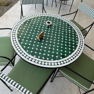 Könnte beinhalten: Garten-Essgruppe mit einem runden Tisch. Der Tisch hat eine grün-weiße Fliesenoberfläche und ist von vier Stühlen umgeben. Die Stühle haben grüne Kissen und schwarze Metallrahmen. Der Tisch hat einen Durchmesser von etwa 100 cm.