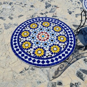 May include: A round mosaic tile table with a blue, yellow, and white geometric pattern. The table has a diameter of approximately 30 inches. The table is outdoors on a stone patio.