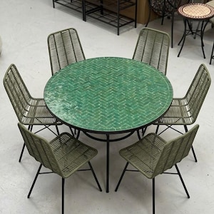 Puede incluir: Una mesa de comedor redonda para exteriores con una superficie de azulejos verdes, rodeada de seis sillas a juego. Las sillas tienen respaldos y asientos tejidos, con patas de metal negro. La mesa y las sillas están dispuestas sobre un suelo claro.