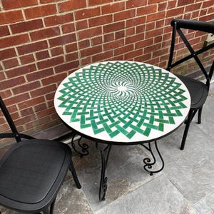 May include: A round outdoor table with a white surface featuring a green geometric spiral design. The table has a black metal frame and is flanked by two black chairs. The table is approximately 76 cm in diameter.