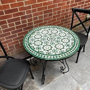 May include: A round outdoor bistro table with a mosaic tile top in shades of green and white. The table has a black metal frame with decorative scrollwork and is paired with two black chairs. The table is approximately 76 cm in diameter.