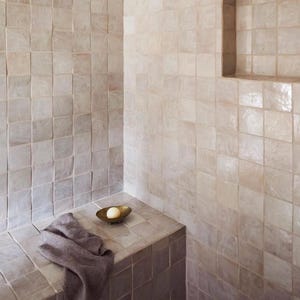 Puede incluir: Un baño con baldosas cuadradas de color gris claro que cubren las paredes y el banco. Un pequeño cuenco dorado contiene una pastilla de jabón. Una toalla gris está sobre el banco. Una estantería empotrada está integrada en la pared.