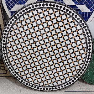 May include: A circular mosaic table top with a geometric design. The pattern includes a grid of white squares, black squares, and orange accents. The edge is decorated with black diamonds and squares. The table top is likely made of ceramic tiles.