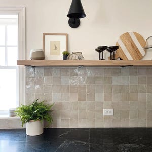 May include: A modern kitchen interior featuring a black countertop, light-colored cabinets, and a backsplash of square tiles. A wooden shelf displays decor, including a framed print, a small plant, and glassware. A black wall sconce and a potted fern add to the aesthetic.
