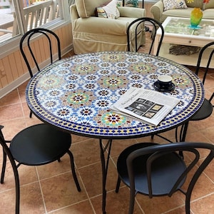 May include: A round mosaic tile table with a black metal frame, surrounded by four black chairs. A newspaper and a cup and saucer sit on the table. The mosaic design features blue, green, and yellow patterns.