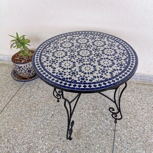 Op de afbeelding: Ronde mozaïek tegel tafel met een donkerblauw en wit geometrisch patroon. De tafel heeft een zwart metalen frame met decoratieve, gebogen poten. Een potplant met groene bladeren staat in de buurt.