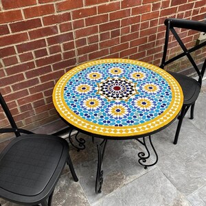 Op de afbeelding: Een ronde buitentafel met een mozaïektegelblad met een geometrisch patroon in blauw, geel en wit. De tafel heeft een zwart metalen frame en wordt geflankeerd door twee zwarte stoelen. De diameter van de tafel is ongeveer 76 cm.