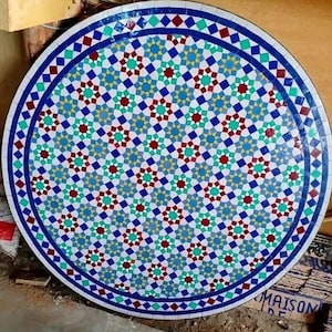 May include: Round mosaic table top with a detailed geometric pattern. The design features a central field of white tiles with teal, red, and yellow star-like shapes. A blue border with diamond and square accents surrounds the central design. The table top is a decorative piece.