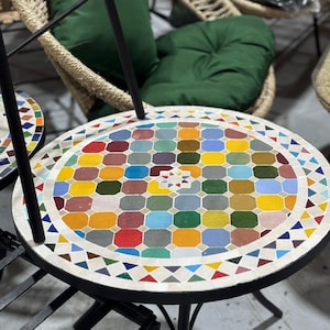 Op de afbeelding: Een ronde mozaïek tegel tafel met een zwart metalen frame. Het tafelblad heeft een kleurrijk geometrisch patroon van vierkanten en driehoeken in verschillende kleuren, waaronder rood, geel, blauw en groen. De tafel maakt deel uit van een patioset.